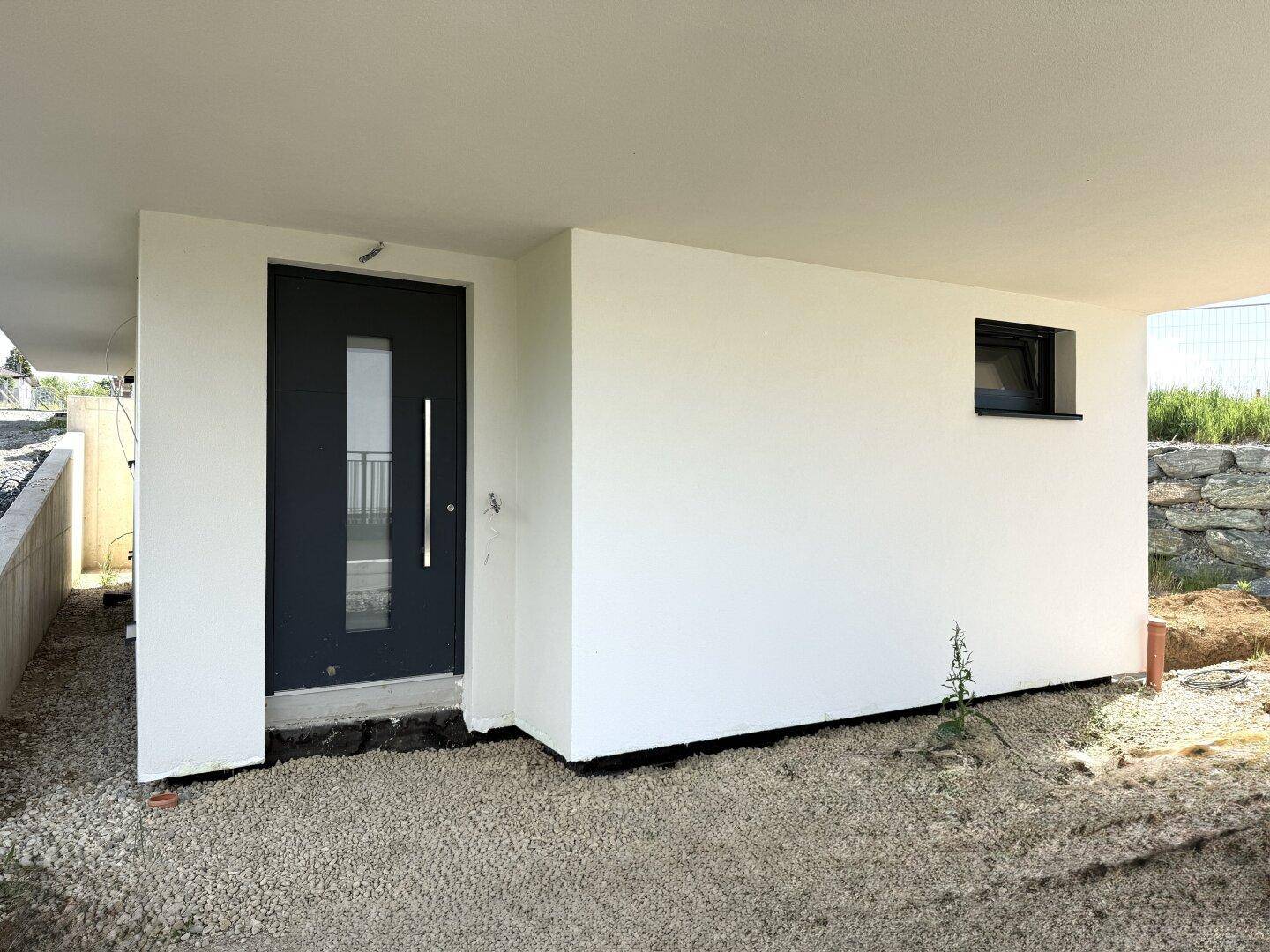 Eingangsbereich eines modernen weißen Hauses mit dunkler Haustür und kleinem Fenster.