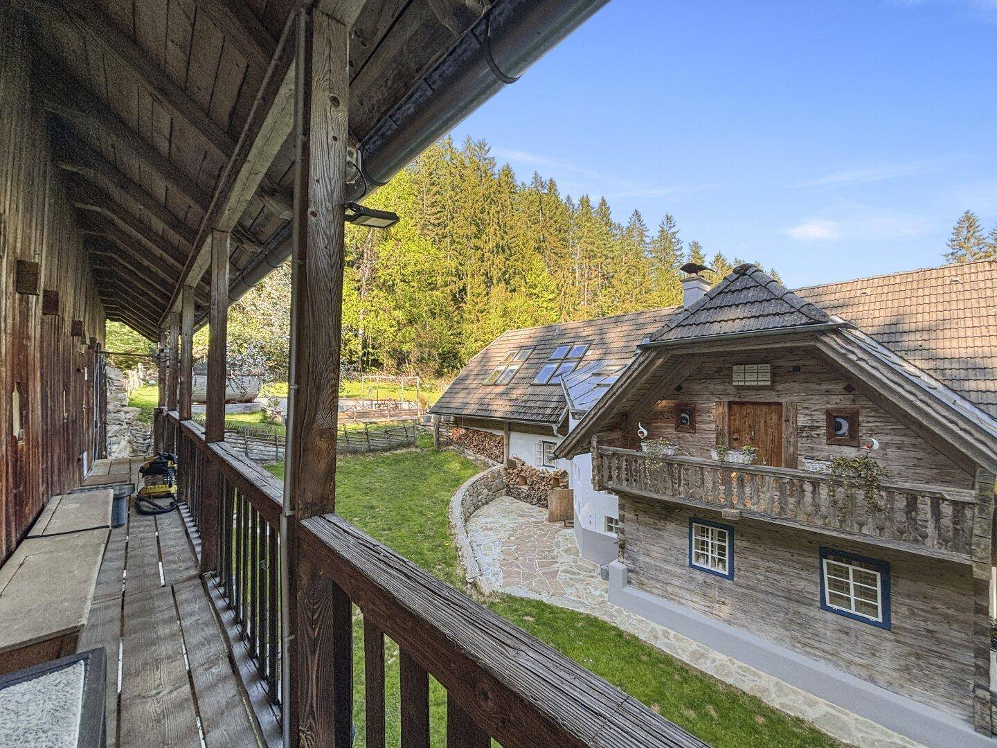 Langer Holzbalkon mit Blick auf weitere Gebäude und den umliegenden Wald in sonniger Lage.