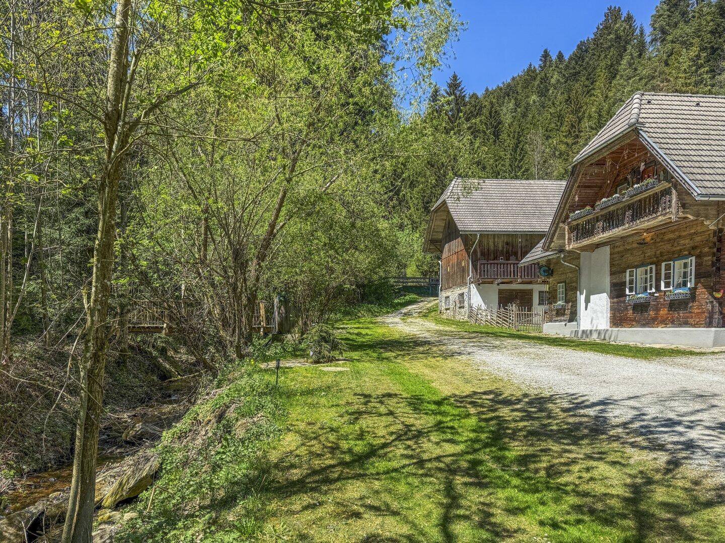 Zufahrt zu den rustikalen Gebäuden, gesäumt von einem Bach und dichtem Wald in ländlicher Umgebung.