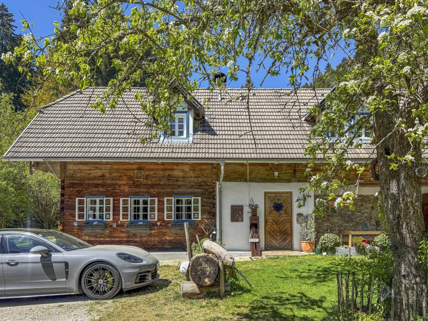 Vorderansicht des traditionellen Holzhauses mit geparktem Auto und blühenden Bäumen im Frühling.