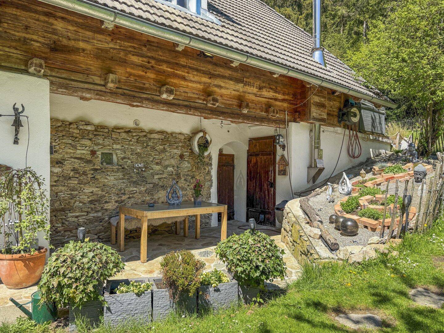 Gemütliche Steinterrasse mit Holztisch und Pflanzen, angrenzend an die rustikale Hausfassade.