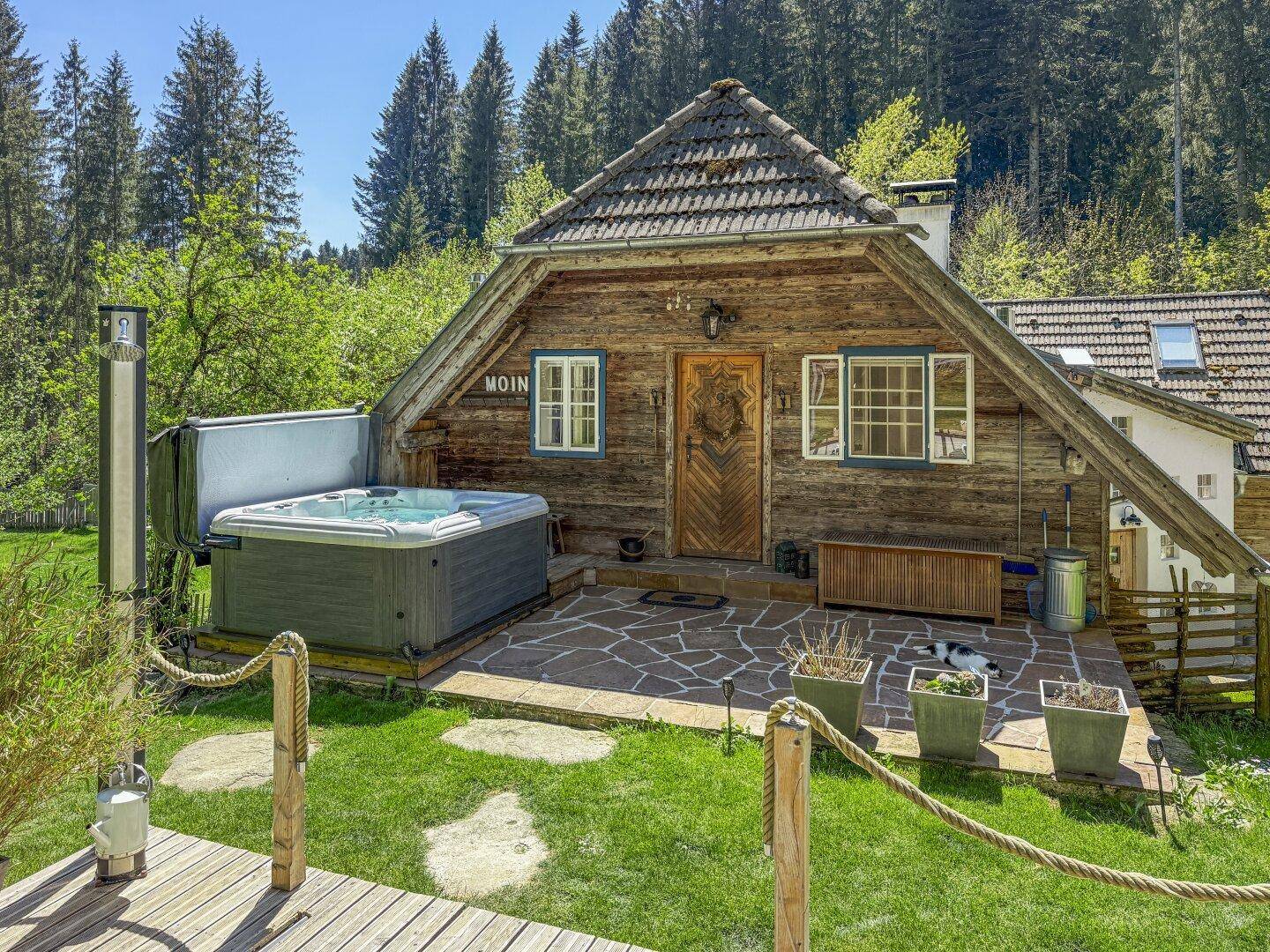 Rustikales Holzhaus mit Whirlpool auf einer Steinterrasse, umgeben von grüner Natur und Wald.