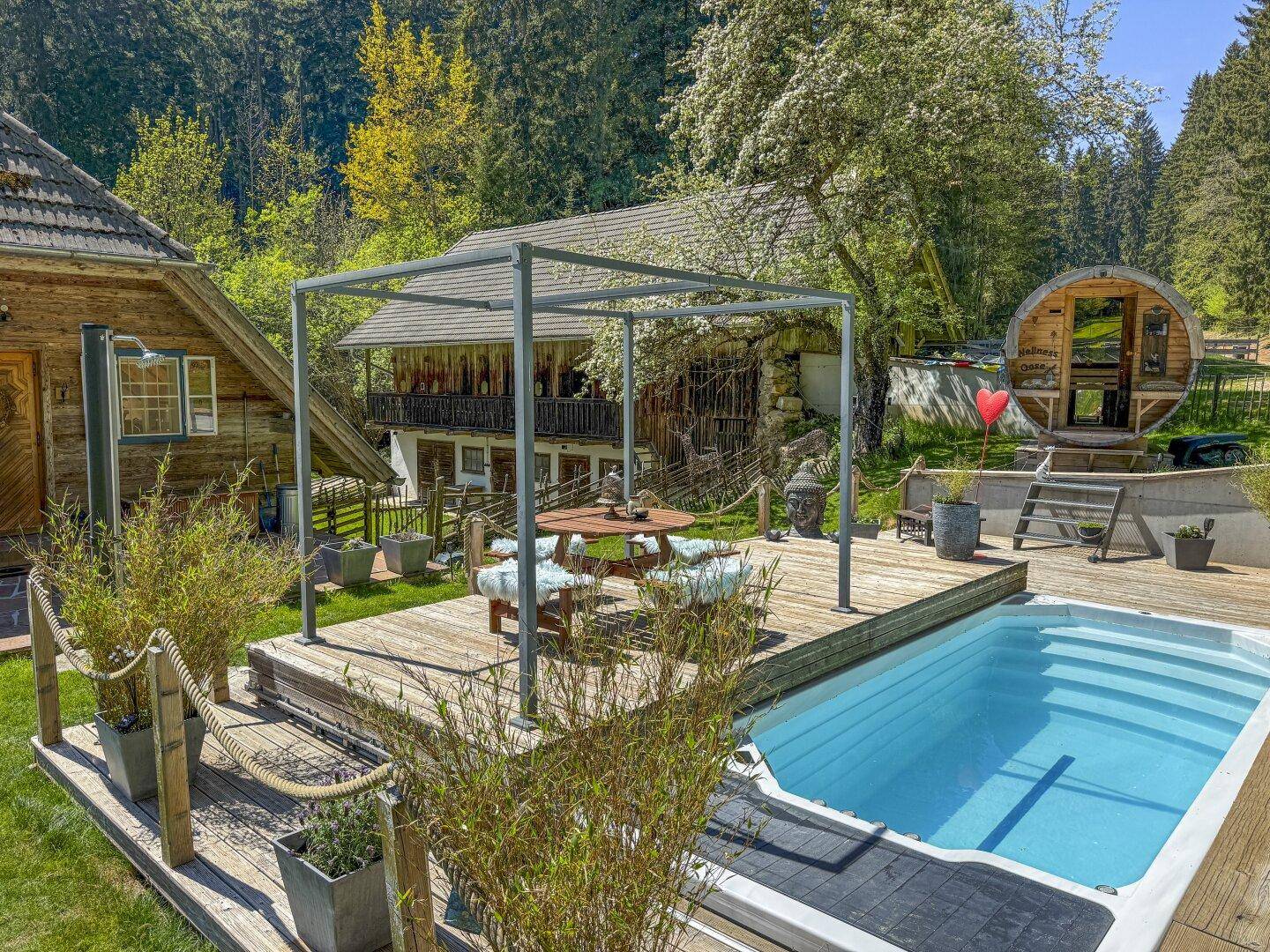 Außenbereich mit Pool, Holzdeck, Pergola und einer Fasssauna im Hintergrund, umgeben von Natur.
