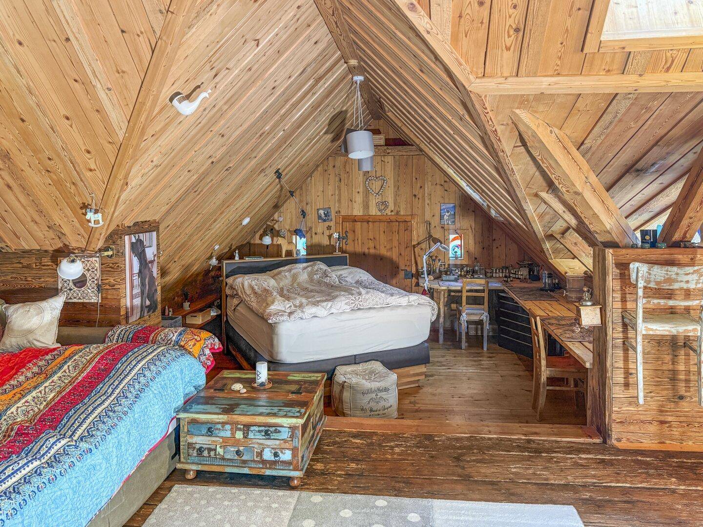 Weitläufiges Dachzimmer mit Holzbalken, gemütlichem Bett und einem integrierten Arbeitsbereich unter den Schrägen.
