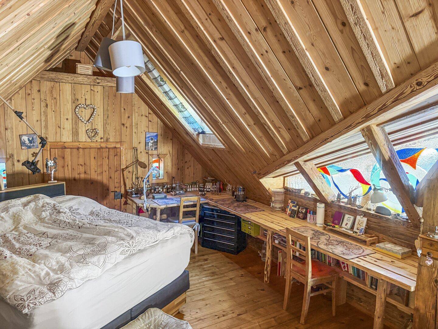 Helles Dachzimmer mit Holzwänden, schrägen Decken, einem Bett und einem langen Schreibtischbereich.