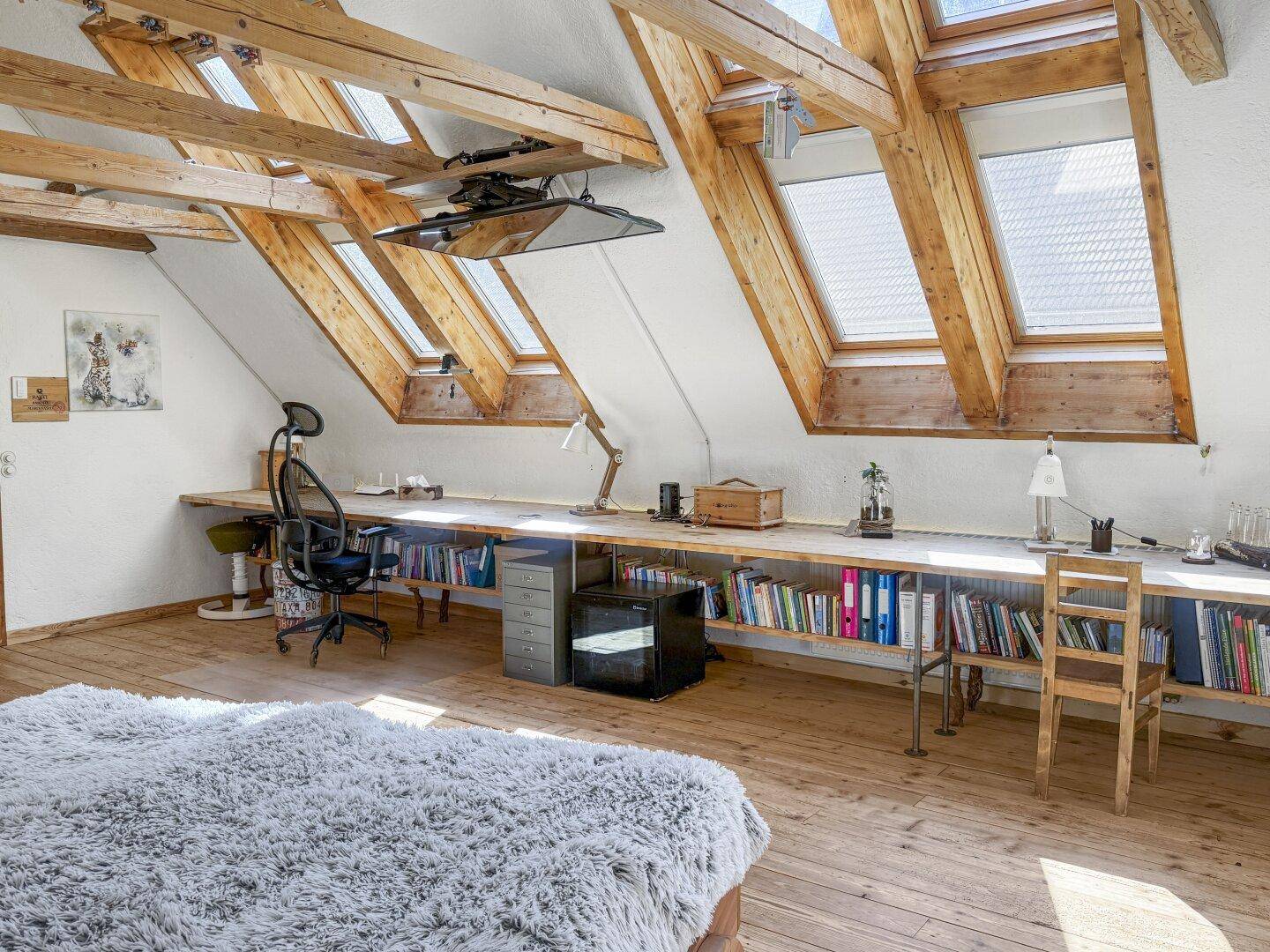 Helles Schlafzimmer im Dachgeschoss mit Holzbalken, Dachfenstern und integriertem Arbeitsbereich.