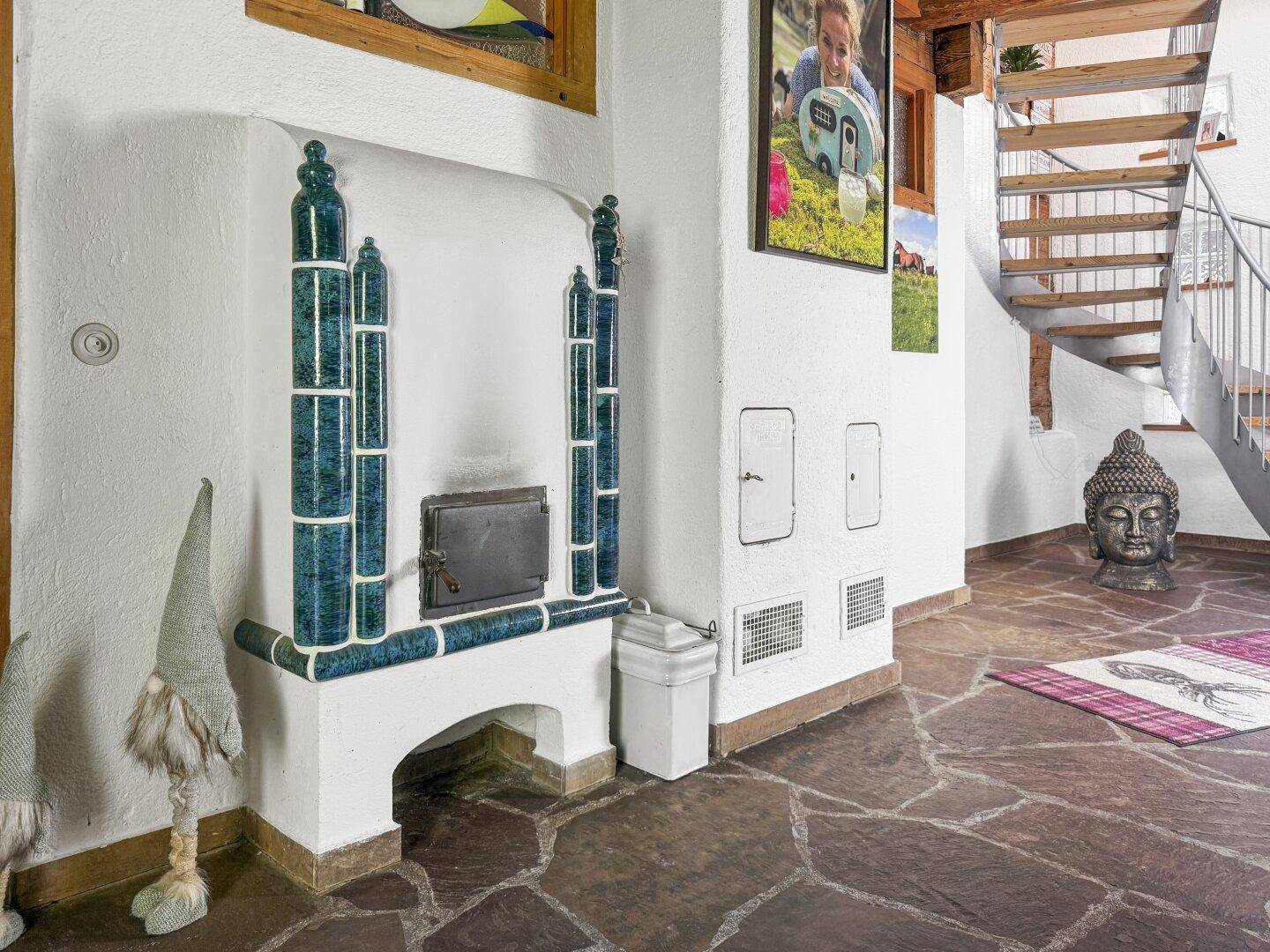 Eingangsbereich mit traditionellem Kachelofen, Steinfliesenboden und offener Holztreppe.