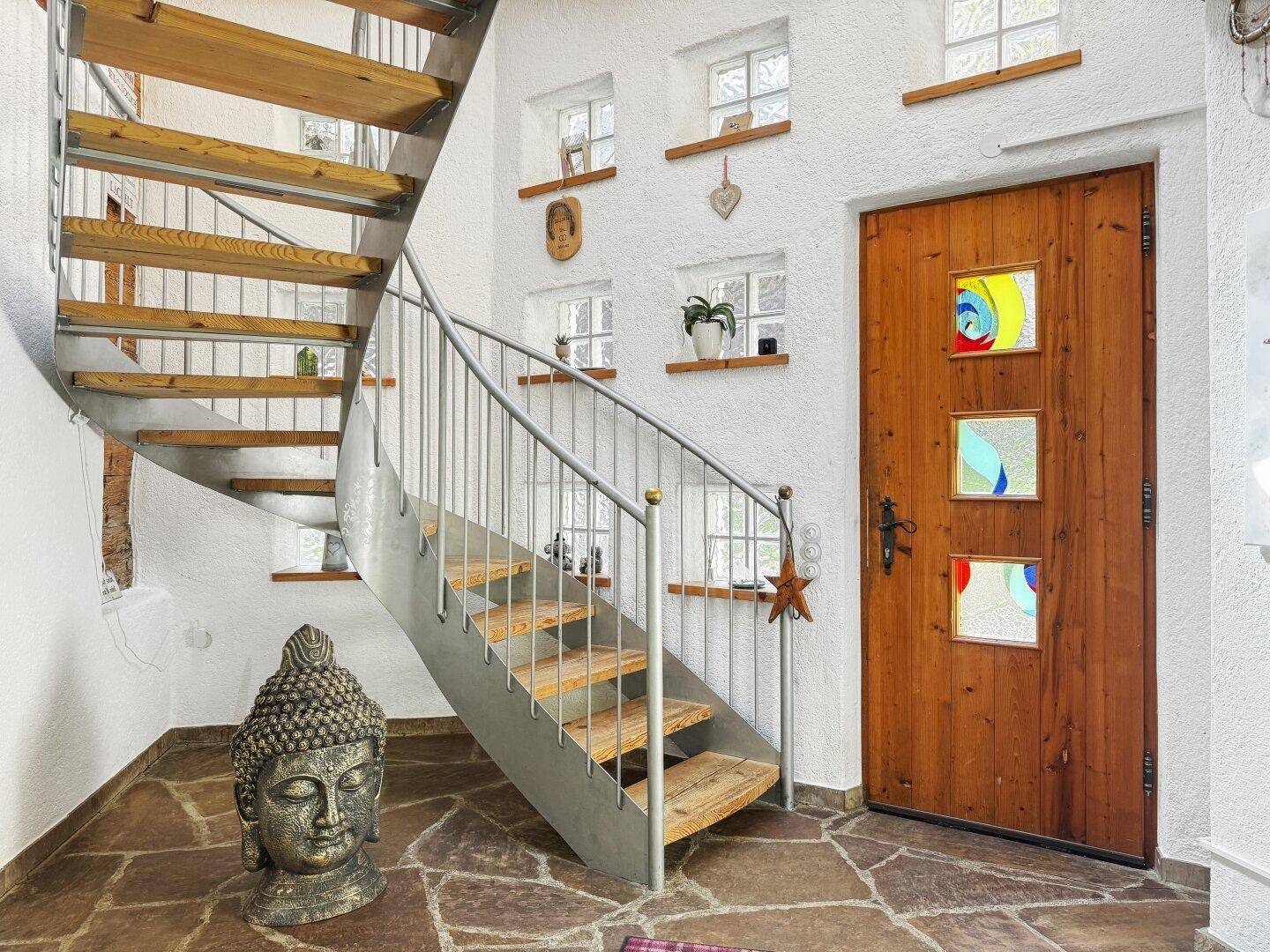 Heller Eingangsbereich mit Wendeltreppe aus Holz und Metall, Steinfliesenboden und Holztür.