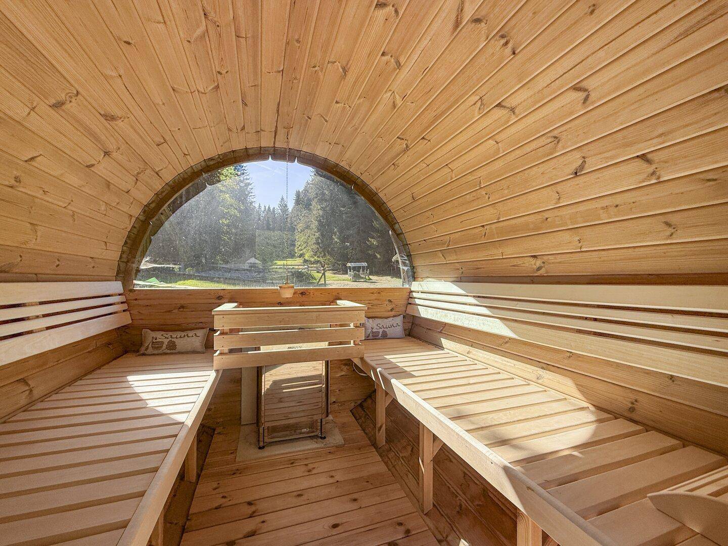 Innenansicht der Holzfasssauna mit Sitzbänken und einem Fenster mit Blick ins Grüne.