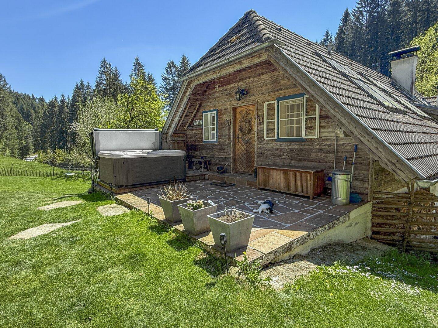 Rustikales Holzhaus mit Whirlpool auf einer Steinterrasse, umgeben von gepflegtem Rasen und Wald.