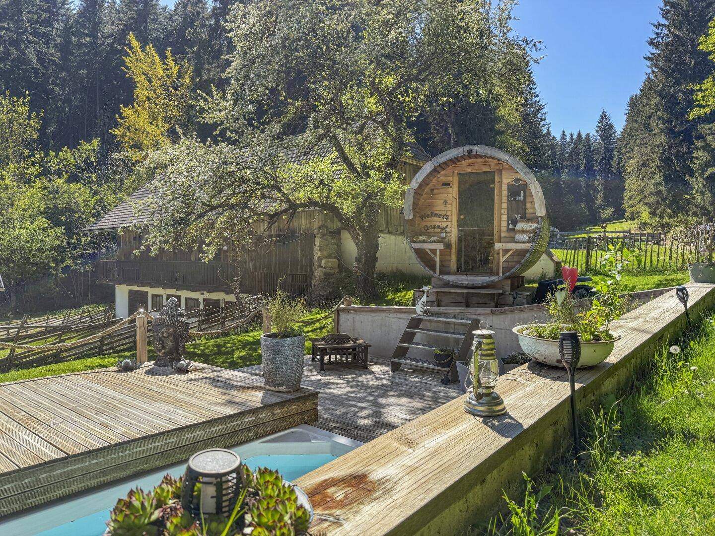 Entspannungsbereich im Garten mit Fasssauna, Pool und Holzterrasse, umgeben von blühenden Bäumen.