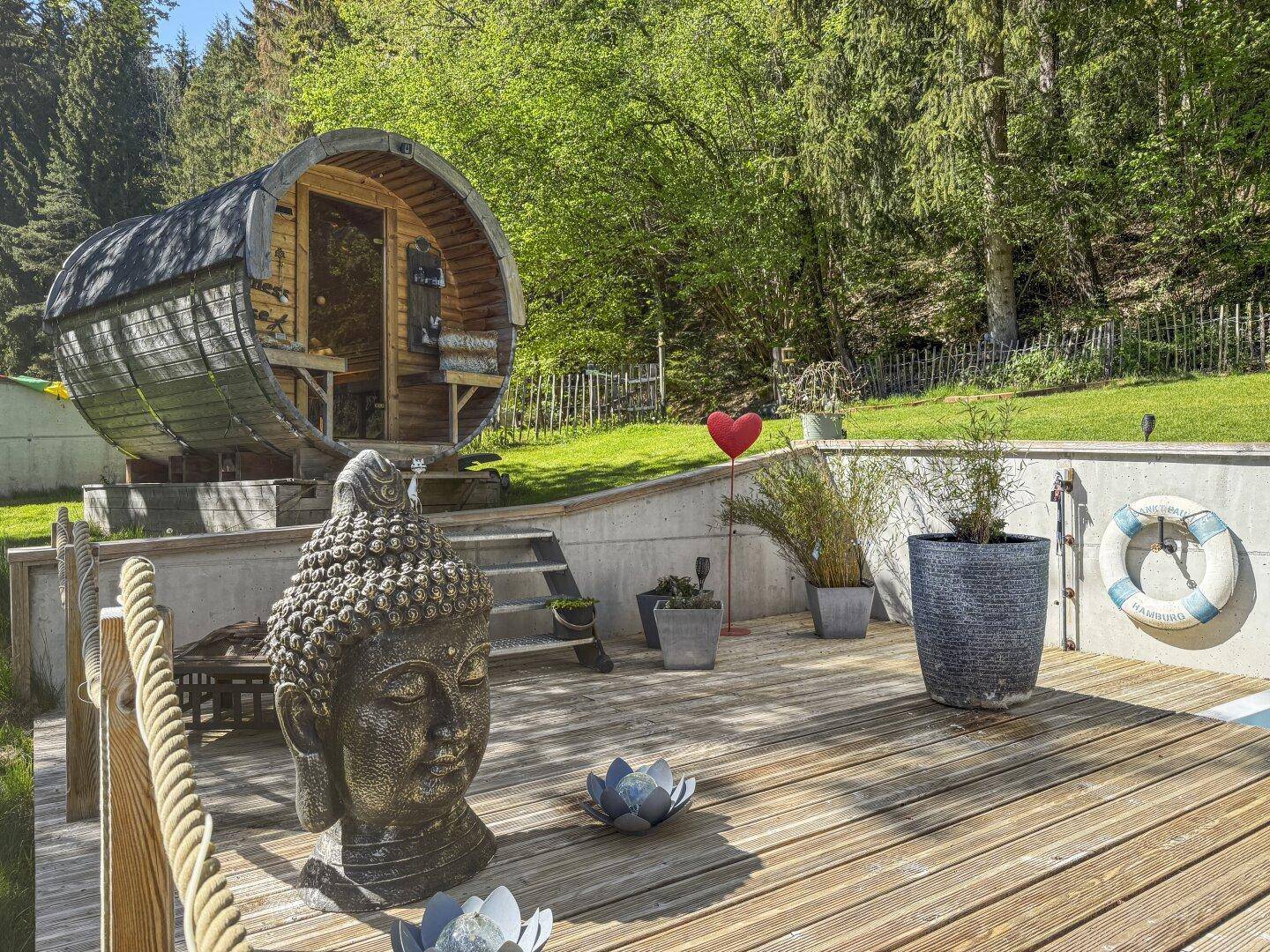 Detailansicht der Poolterrasse mit Buddha-Statue und der Fasssauna im Hintergrund, umgeben von Grün.