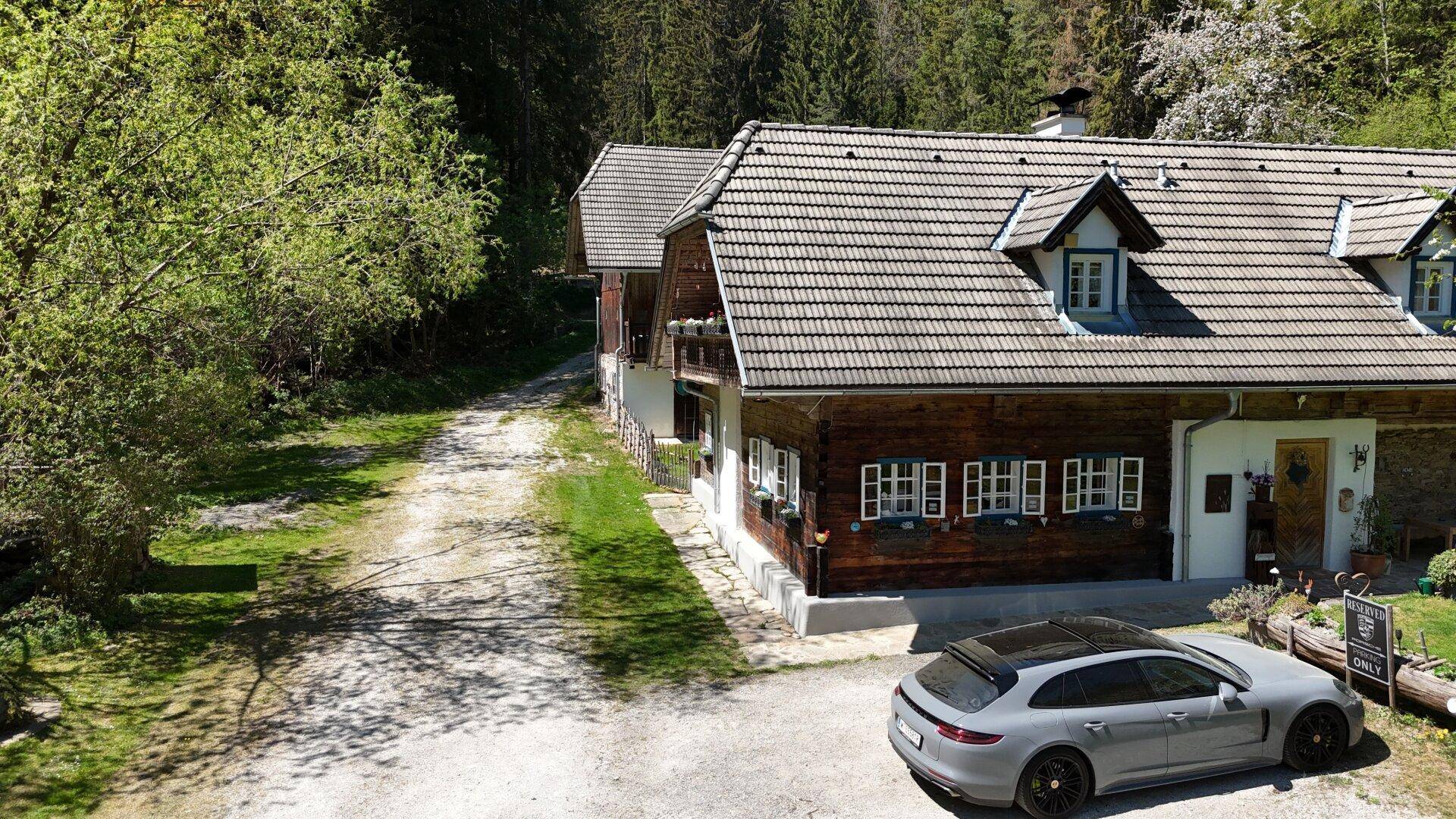 Rustikales Holzhaus mit Satteldach und Kiesauffahrt, umgeben von grüner Natur und Wald.