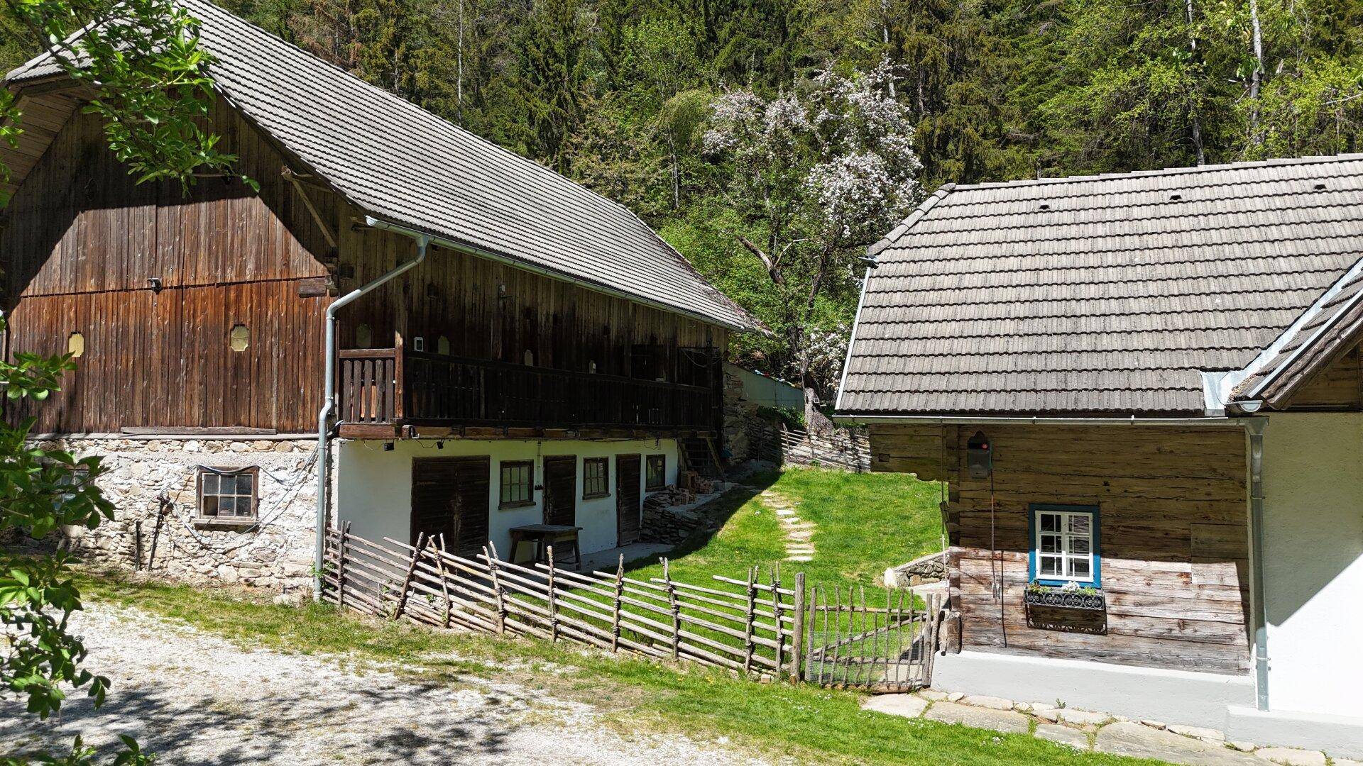 Zwei rustikale Holzhäuser mit traditionellen Dächern, eingebettet in eine grüne Hügellandschaft.