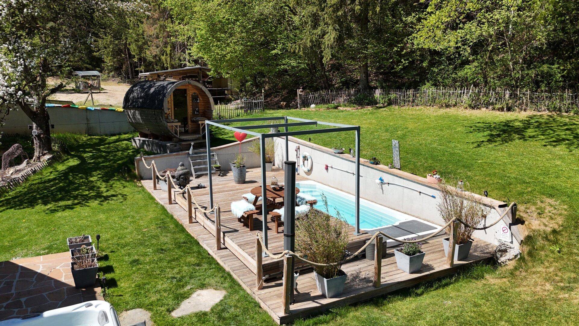 Idyllischer Gartenbereich mit Pool, Holzterrasse, Fasssauna und viel Grün.