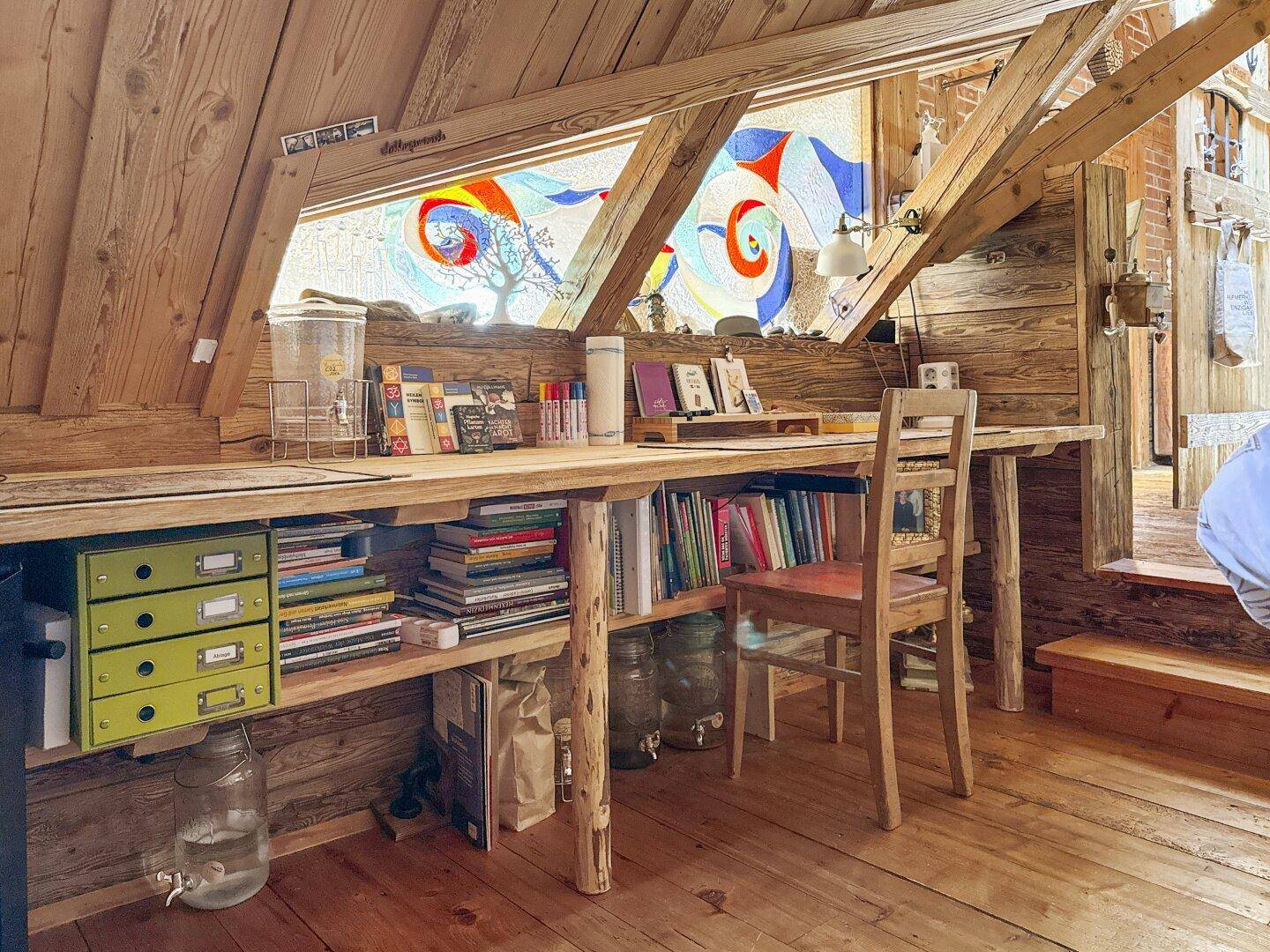 Detailansicht des Arbeitsbereichs im Dachgeschoss mit Holztisch, Büchern und farbigen Fenstern.