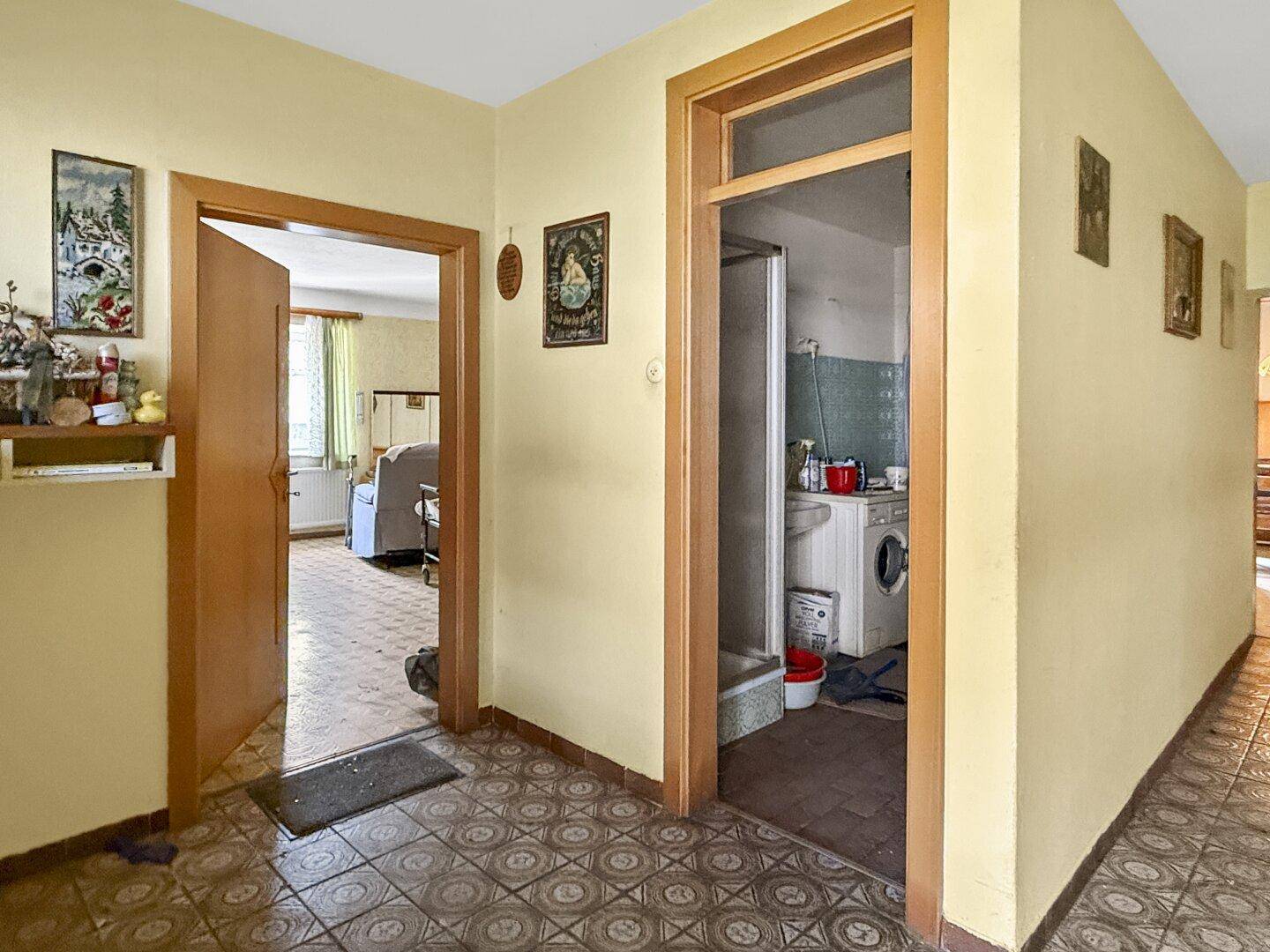 Flur mit gemusterten Bodenfliesen und Holztürrahmen, Blick in ein Zimmer und einen Duschbereich.