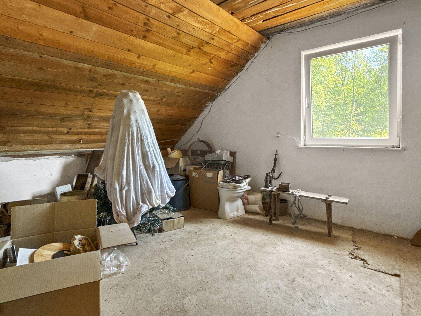 Dachboden mit Holzbalken und einem Fenster, das den Blick ins Grüne freigibt.