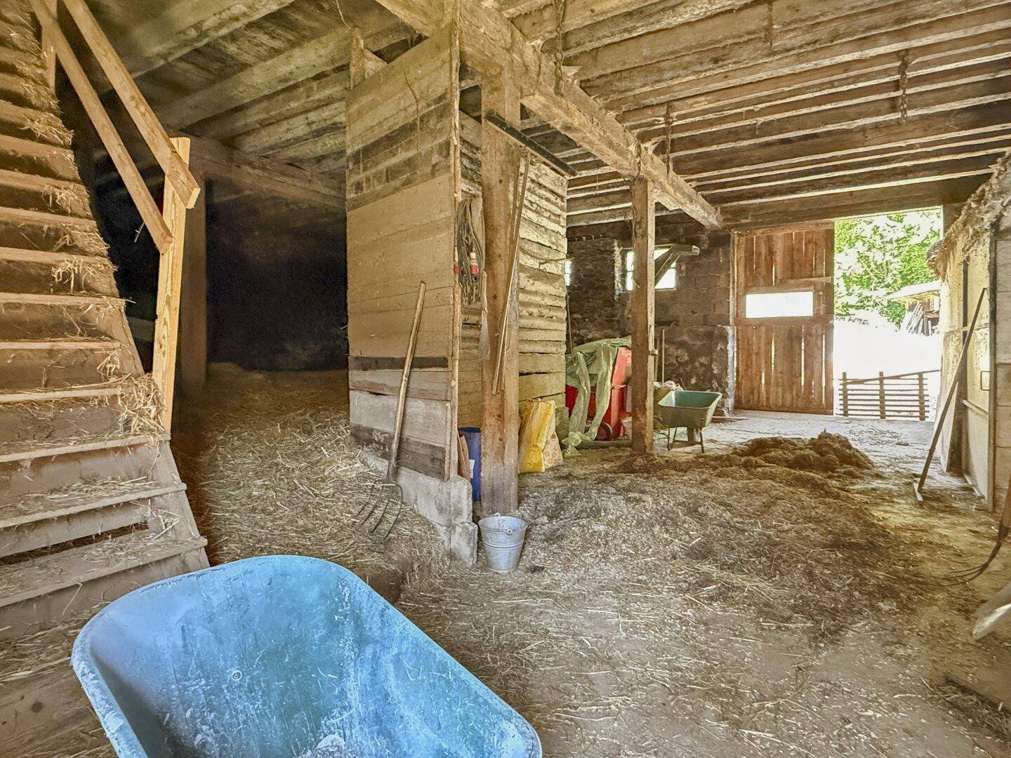 Innenansicht einer Scheune mit Heu, landwirtschaftlichen Geräten und Holzbalken.