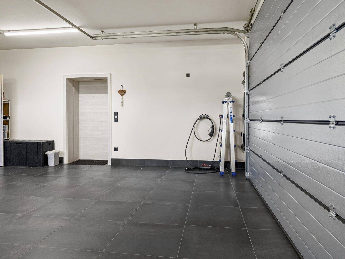 Geräumige Garage mit Fliesenboden, Sektionaltor und einer Ladestation für Elektrofahrzeuge.