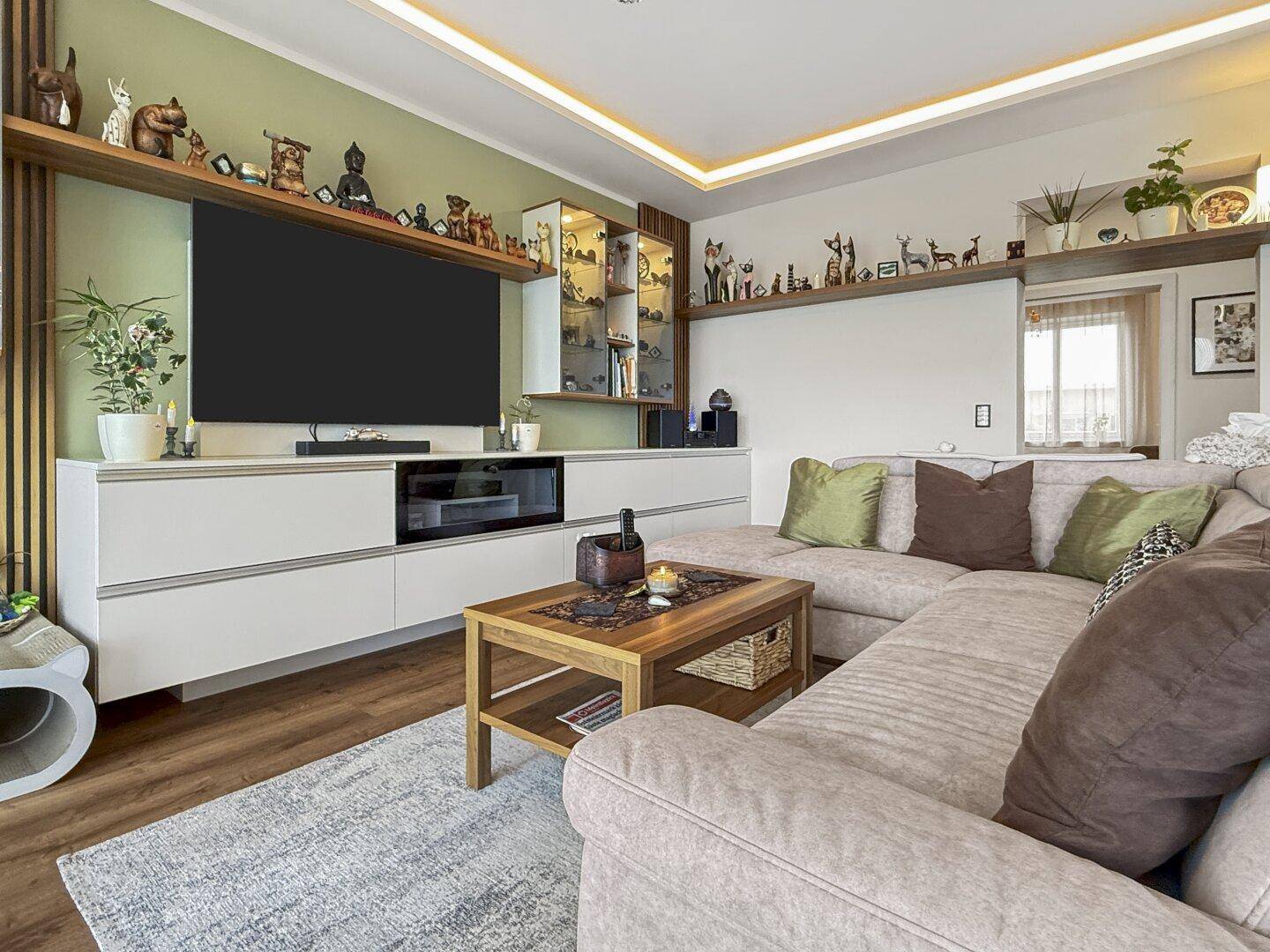 Heller Wohnbereich mit großem Fernseher, Ecksofa und indirekter Deckenbeleuchtung für Ambiente.