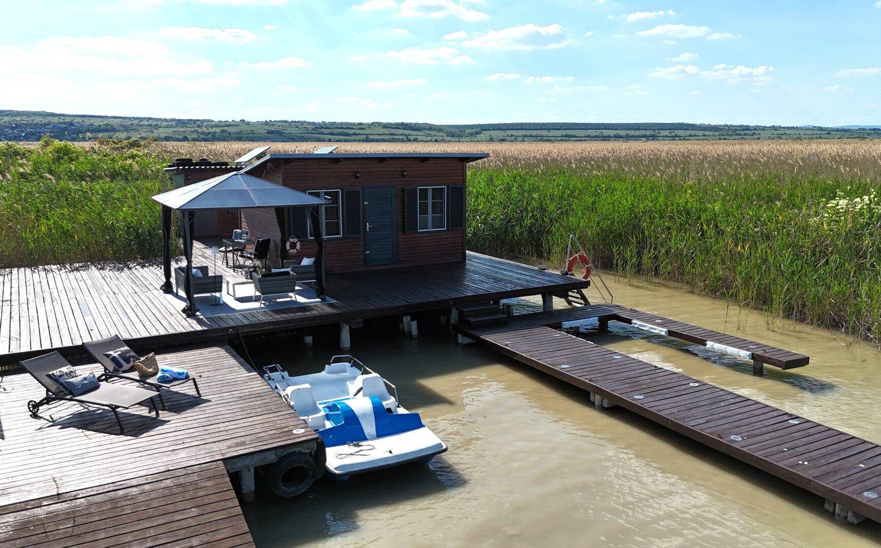 Holzhaus auf einem Steg mit möblierter Terrasse, Sonnenliegen und einem Tretboot am Ufer.