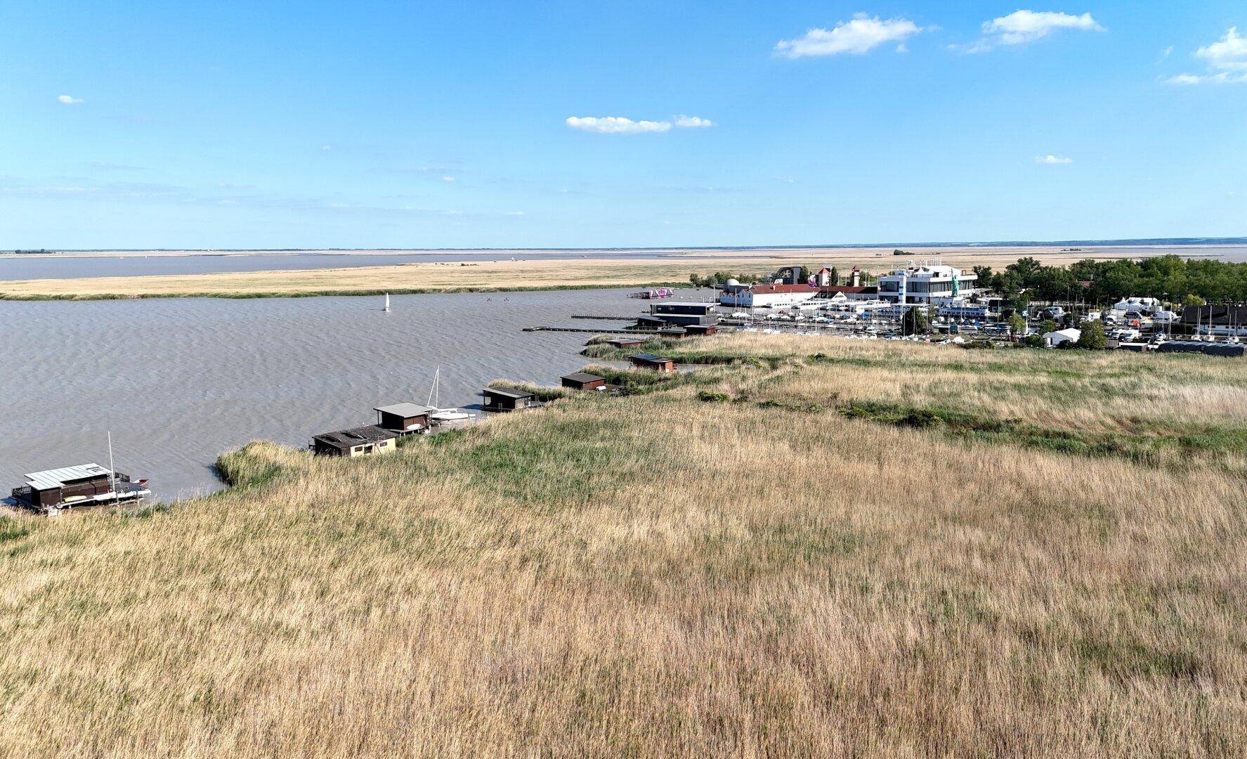 Weitläufiger Blick über den See mit Schilfgürtel und einer entfernten Marina am Horizont.