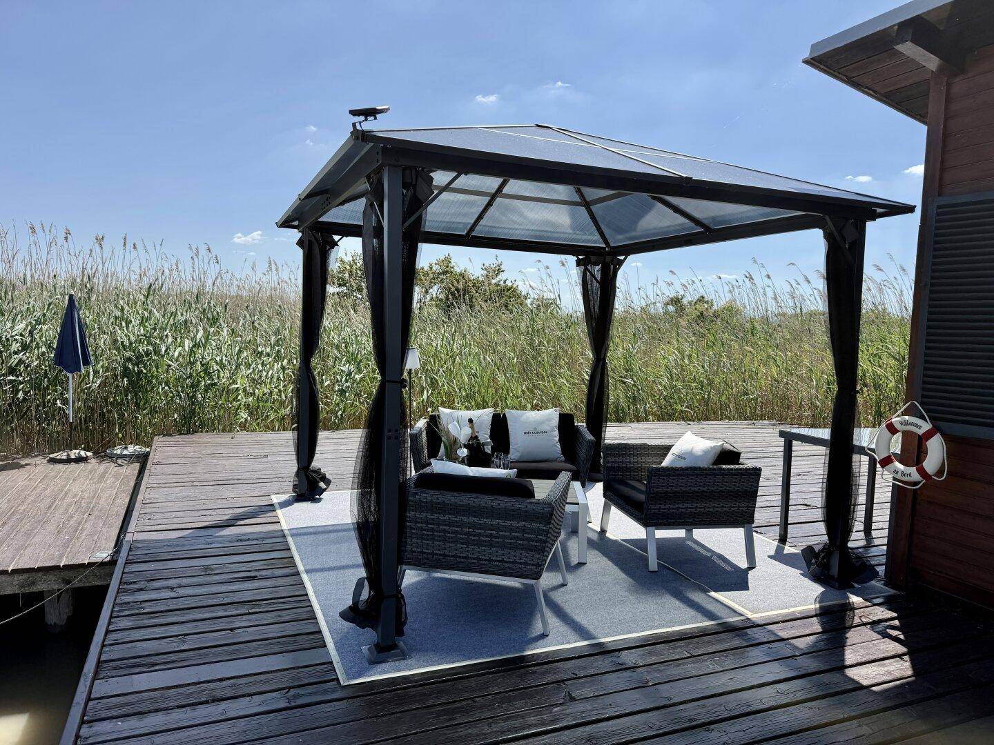 Eleganter Pavillon mit Lounge-Möbeln auf dem Holzsteg, umgeben von üppigem Schilf.