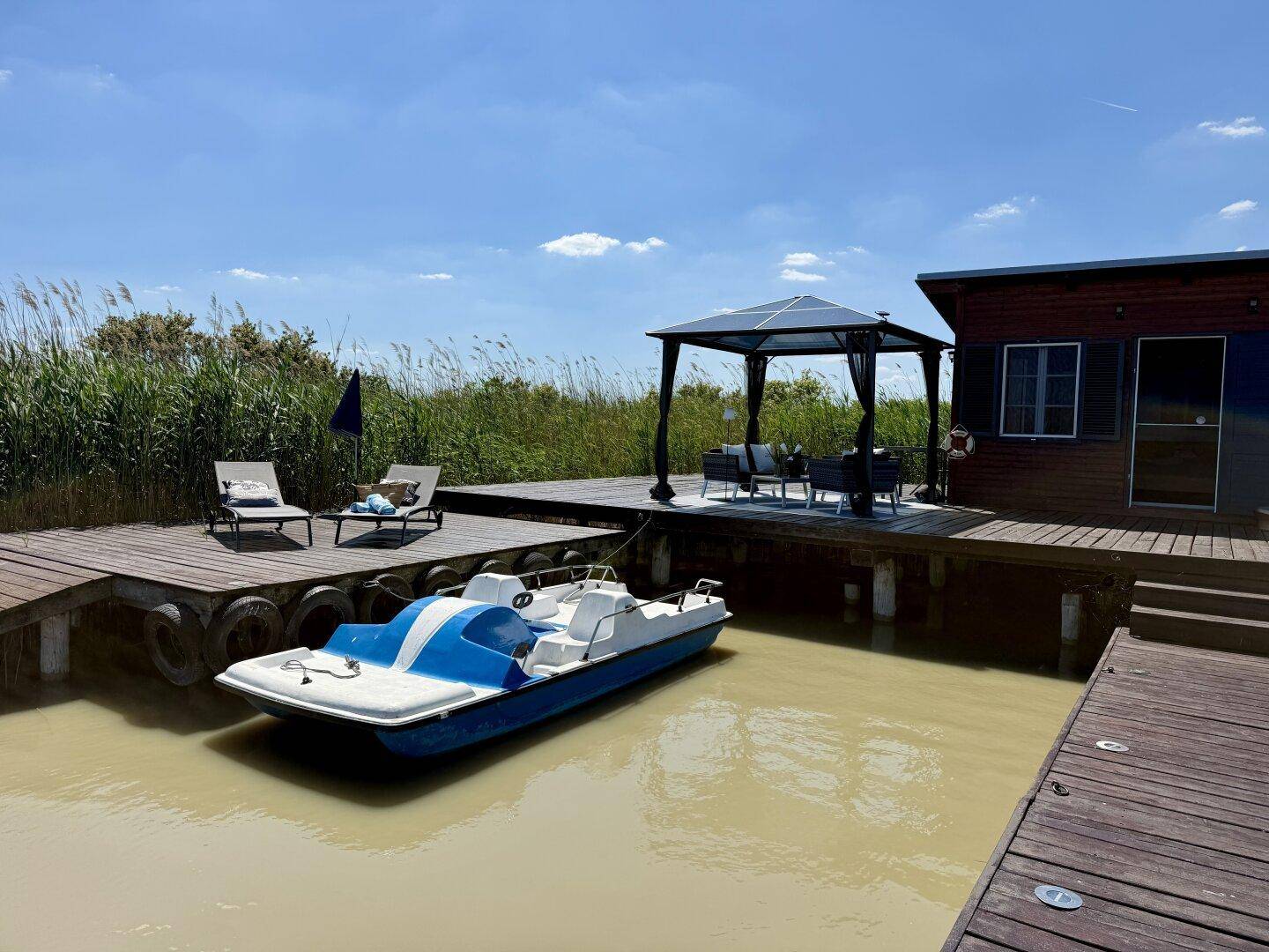 Steg mit Tretboot, Sonnenliegen und einem Pavillon mit Sitzgruppe, umgeben von Schilf.