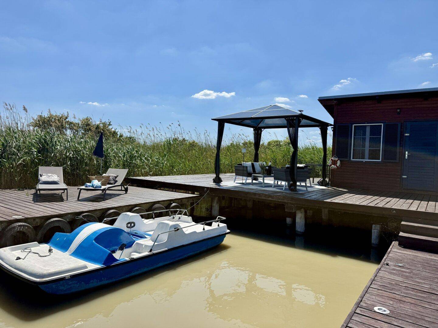 Sonniger Steg mit Tretboot, bequemen Sonnenliegen und einem schattenspendenden Pavillon.