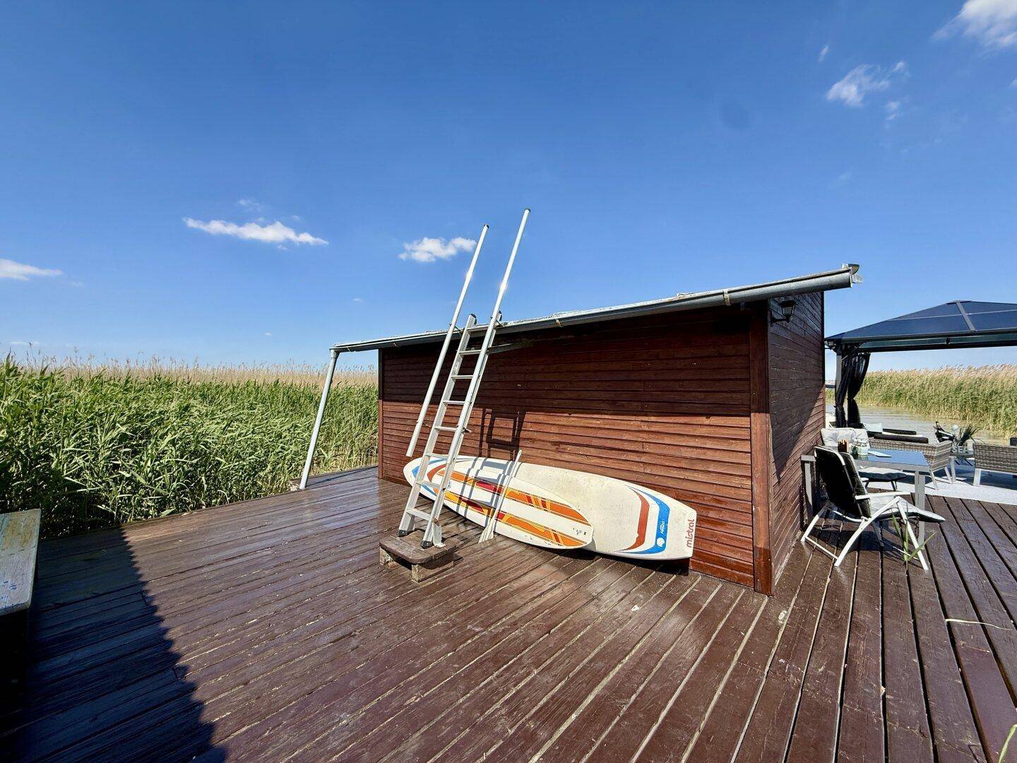 Kleines Holzhaus auf dem Steg mit Leiter und einem Paddleboard, umgeben von Schilf.