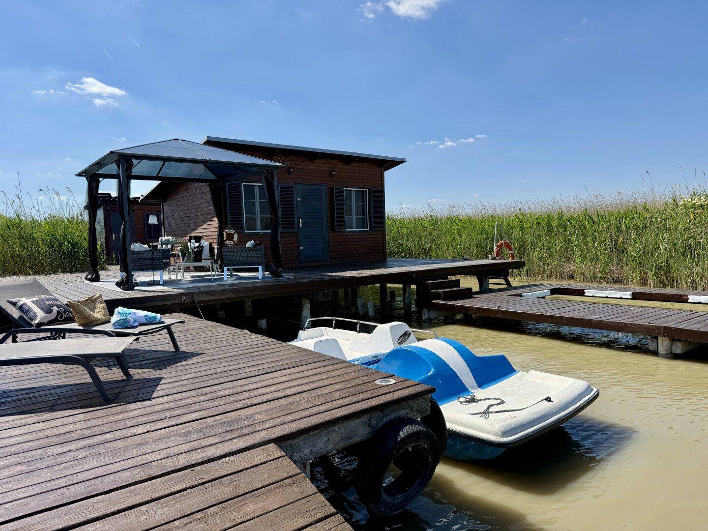 Weitläufiger Holzsteg mit Tretboot, Sonnenliegen, Holzhaus und einem schattenspendenden Pavillon.