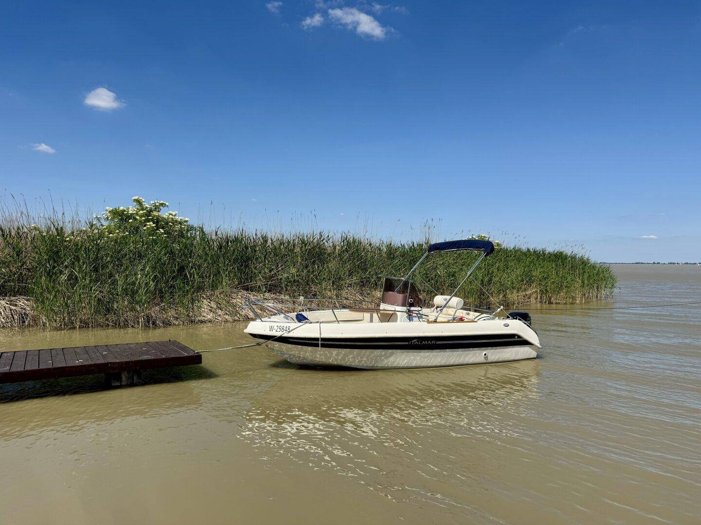 Ein Boot liegt an einem Holzsteg, umgeben von Schilf und dem weiten See unter blauem Himmel.