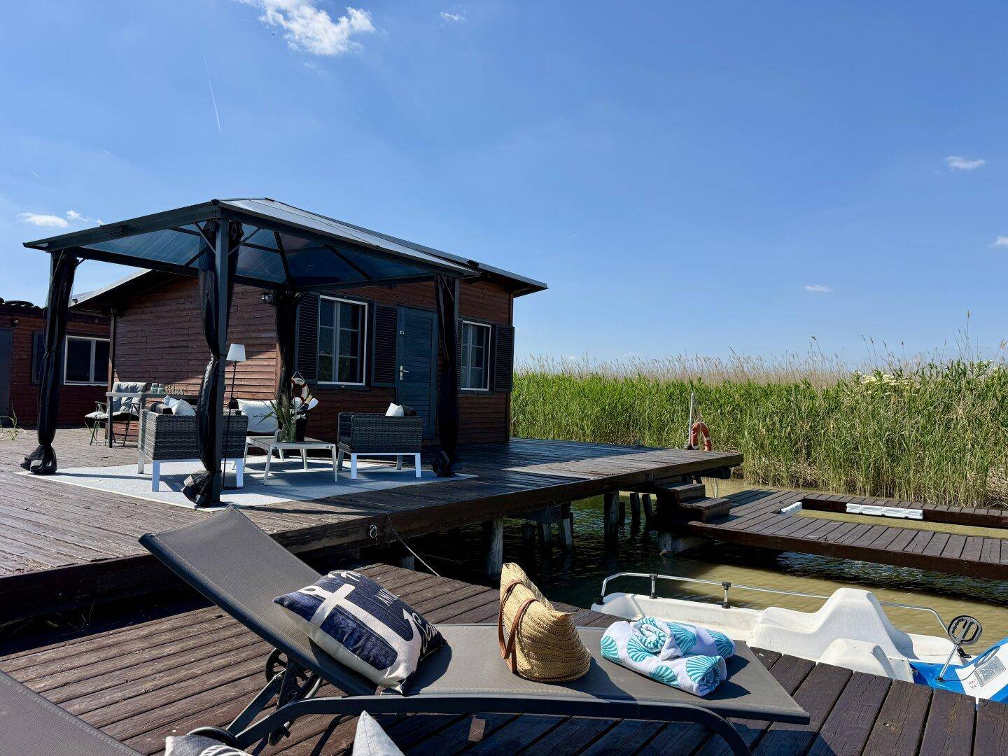 Entspannter Blick von der Sonnenliege auf das Holzhaus und den Pavillon am Wasser.