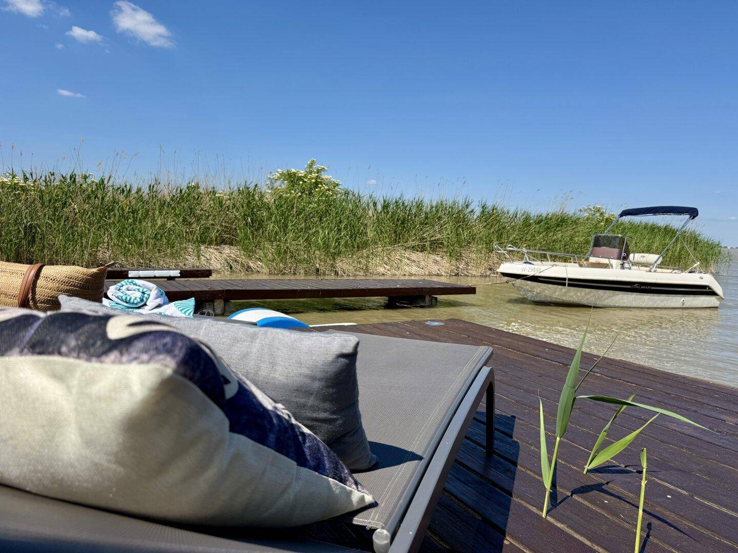 Bequeme Sonnenliege mit Kissen auf dem Steg, mit Blick auf das Wasser und ein Boot.