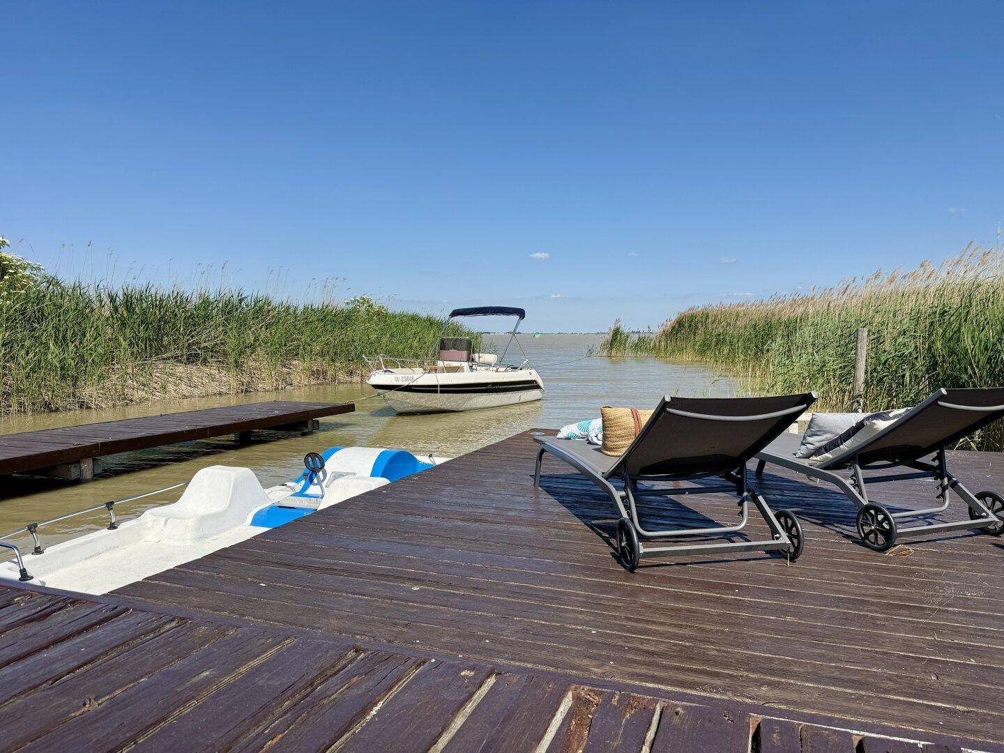 Zwei Sonnenliegen auf dem Holzsteg mit Blick auf das Tretboot und ein Motorboot auf dem See.