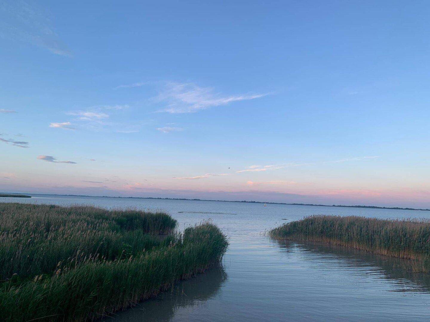 Malerischer Sonnenuntergang über dem See, mit ruhigem Wasser und Schilf im Vordergrund.
