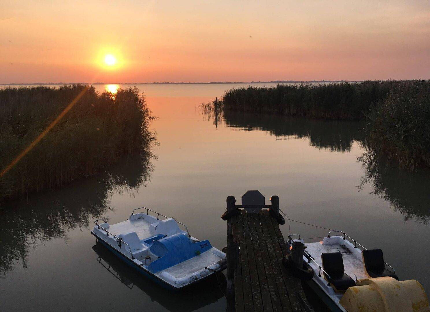 Sonnenuntergang über dem See, mit Booten am Steg und warmem Licht über der Landschaft.