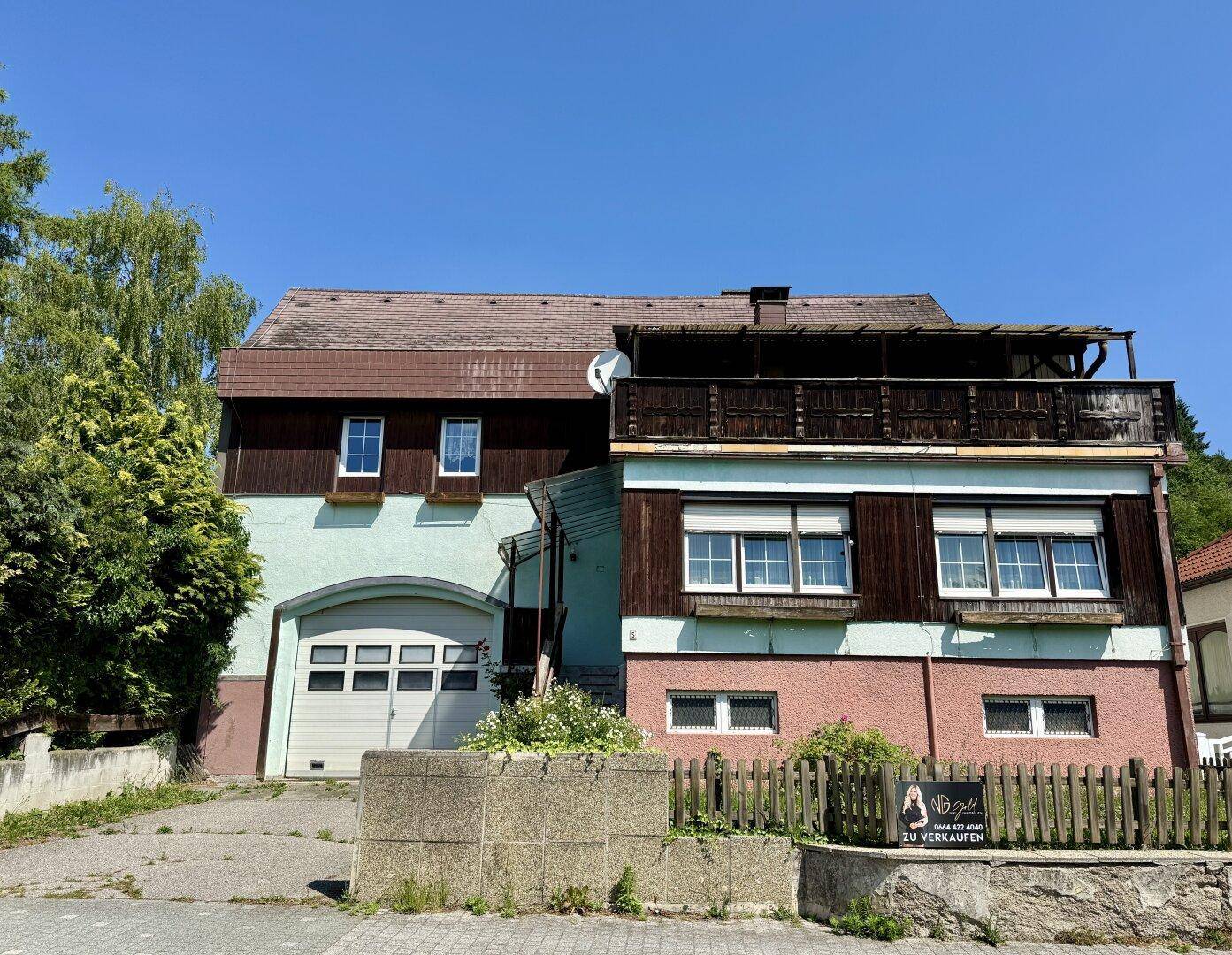 Frontansicht des Hauses mit Garage, Balkon und einem "Zu Verkaufen" Schild im Vorgarten.