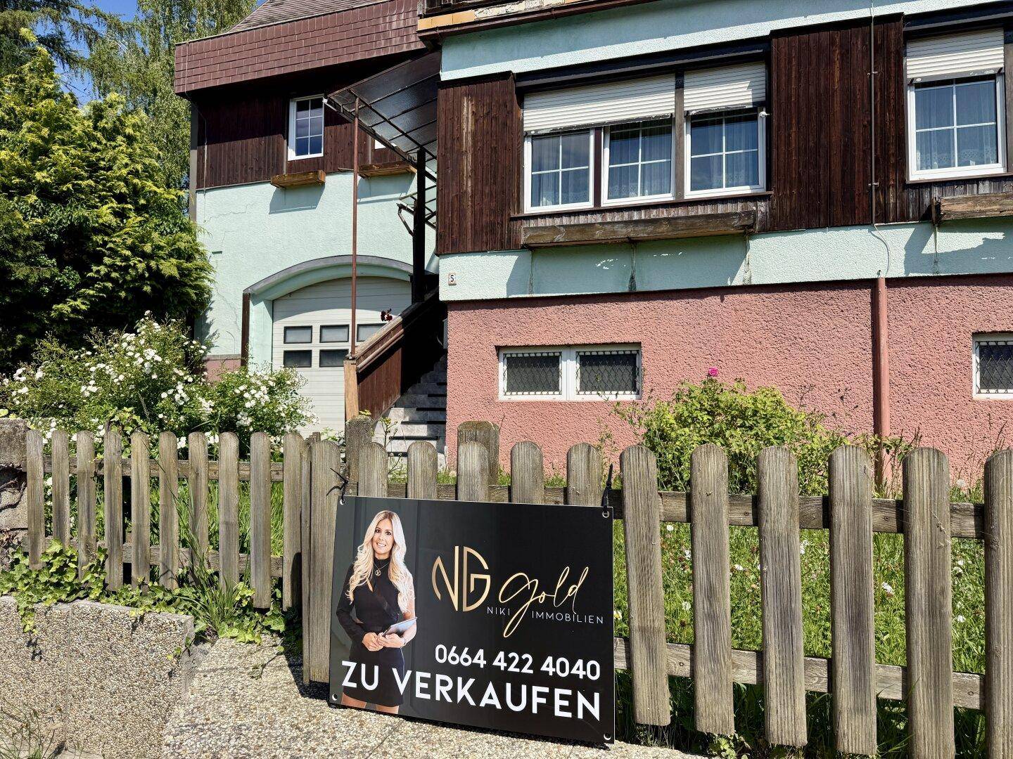 Vorderansicht des Hauses mit einem "Zu Verkaufen" Schild und Holzzaun im Vorgarten.