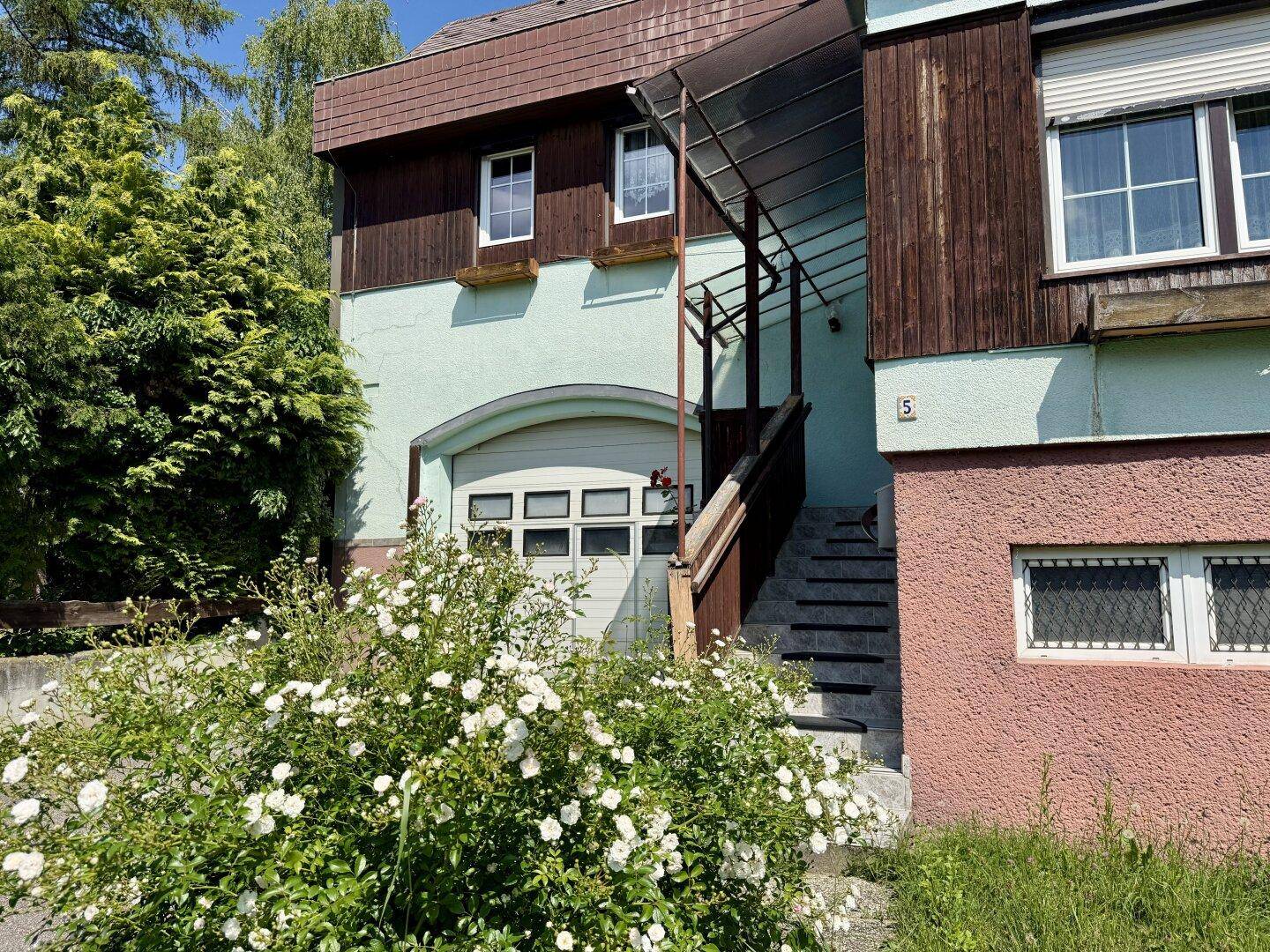 Detailansicht der Hausfront mit Garage, Außentreppe und blühenden Büschen.