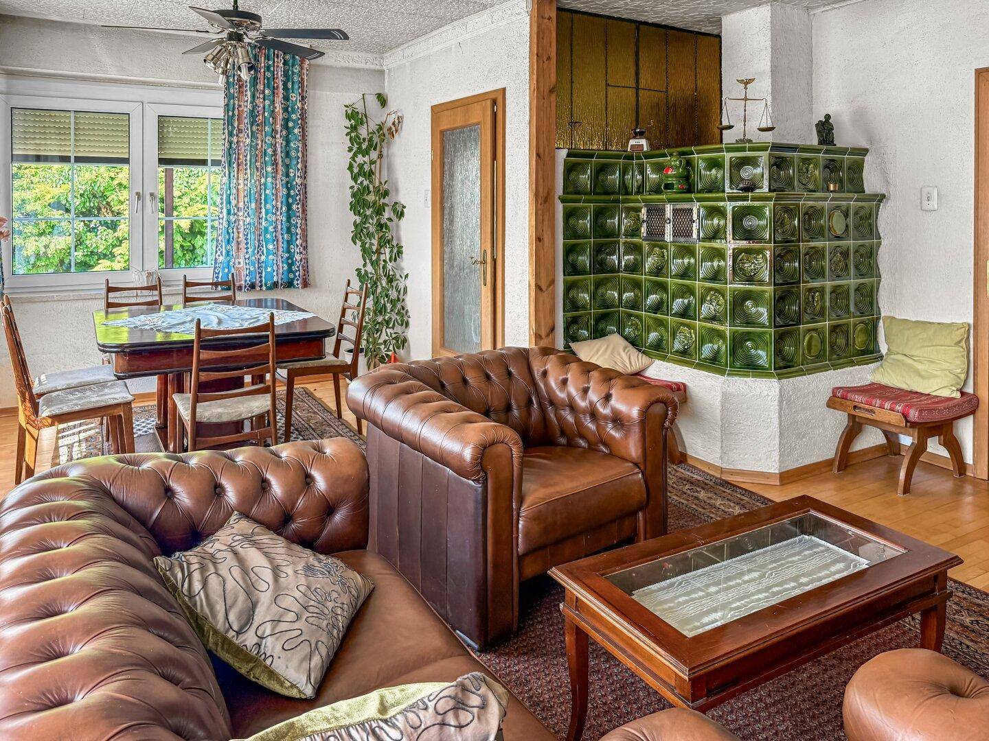 Gemütliches Wohnzimmer mit Ledersofa, Kachelofen und Essbereich am Fenster.
