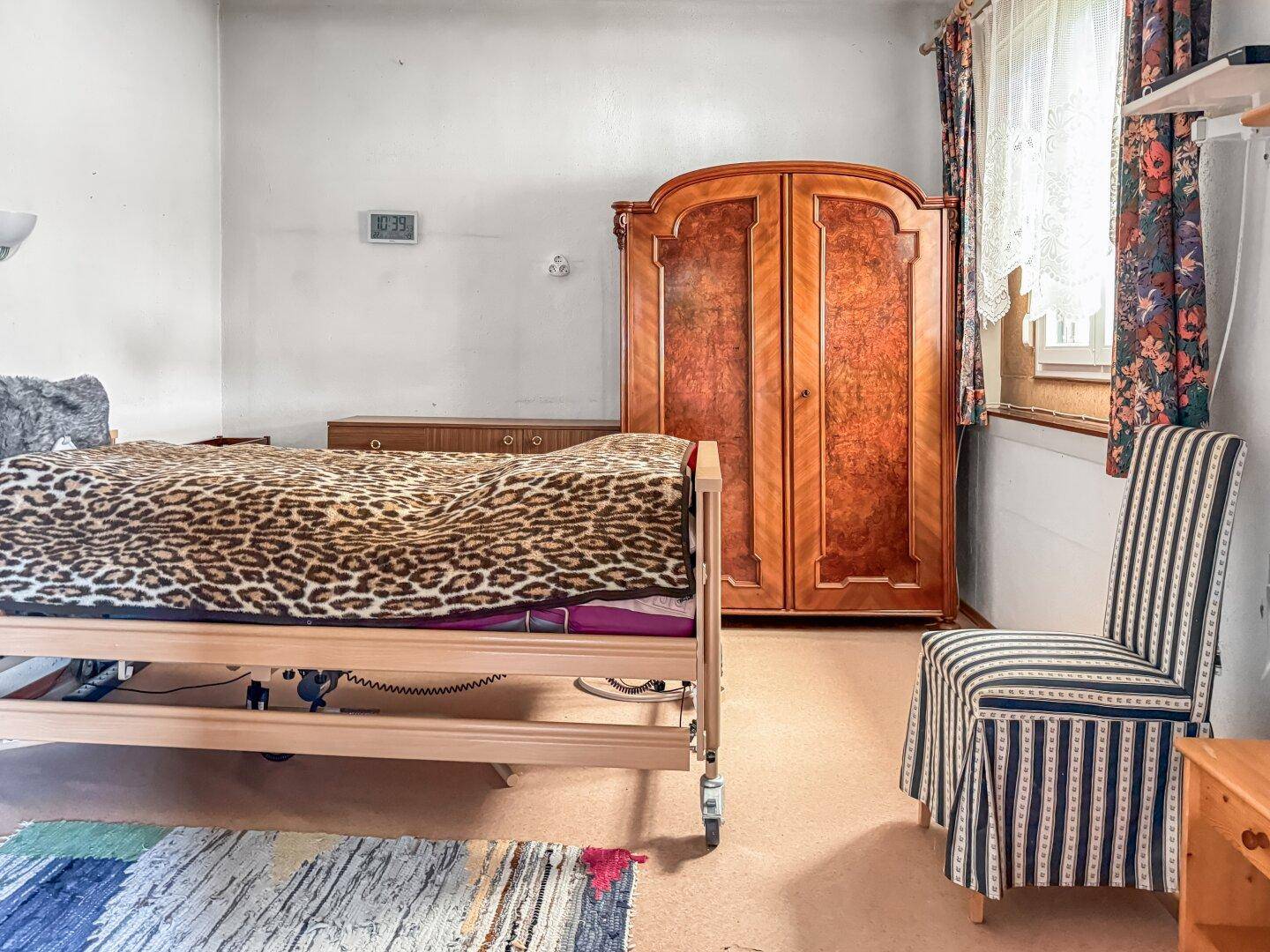 Schlafzimmer mit Pflegebett, Holzschrank und Fenster mit Vorhängen.