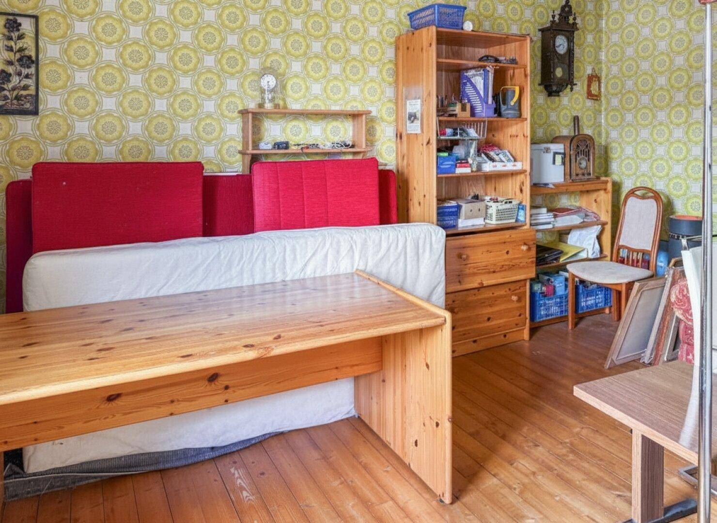 Wohnraum mit ausziehbarem Sofa, Holztisch und Regal, umgeben von einer auffälligen Mustertapete.