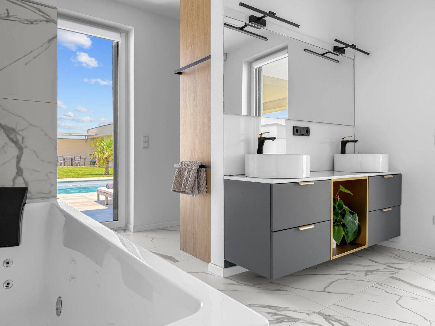Helles Badezimmer mit Whirlpool, Doppelwaschbecken und Blick auf den Außenbereich mit Pool.
