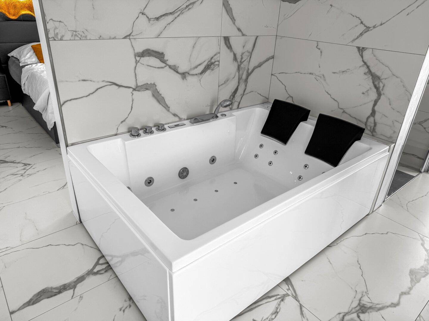 Geräumige Whirlpool-Badewanne mit Marmoroptik-Fliesen und Blick auf das angrenzende Schlafzimmer.