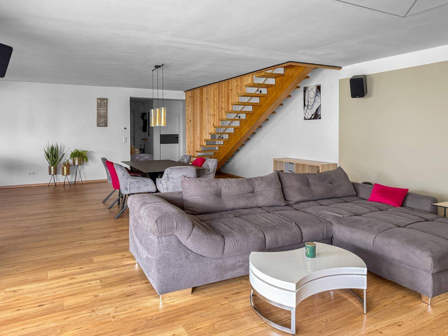 Offener Wohn-Essbereich mit grauem Sofa, Esstisch und eleganter Holztreppe.