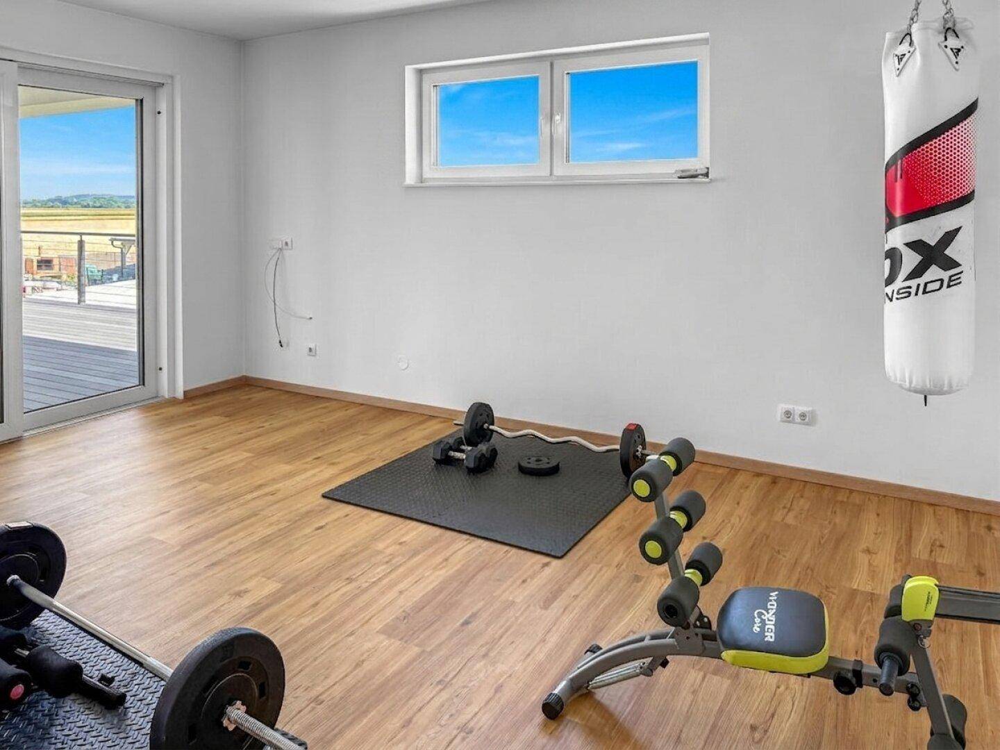 Heller Fitnessraum mit Trainingsgeräten, Hanteln und Fenster mit Blick ins Grüne.