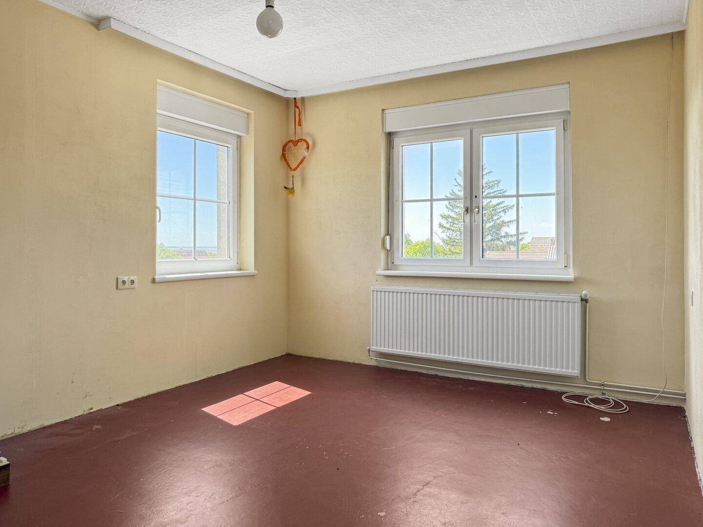 Helles Schlafzimmer mit rotem Boden und zwei Fenstern, die den Raum gut belichten.