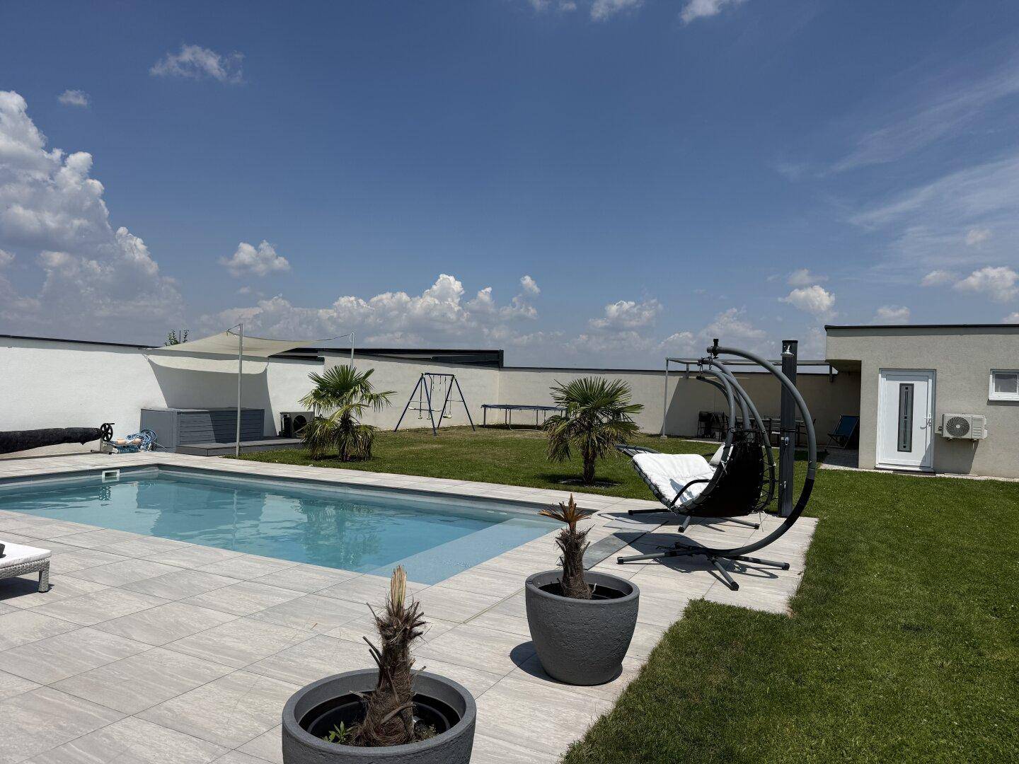 Großer Gartenbereich mit Pool, Liegeflächen, Schaukel und Palmen unter blauem Himmel.
